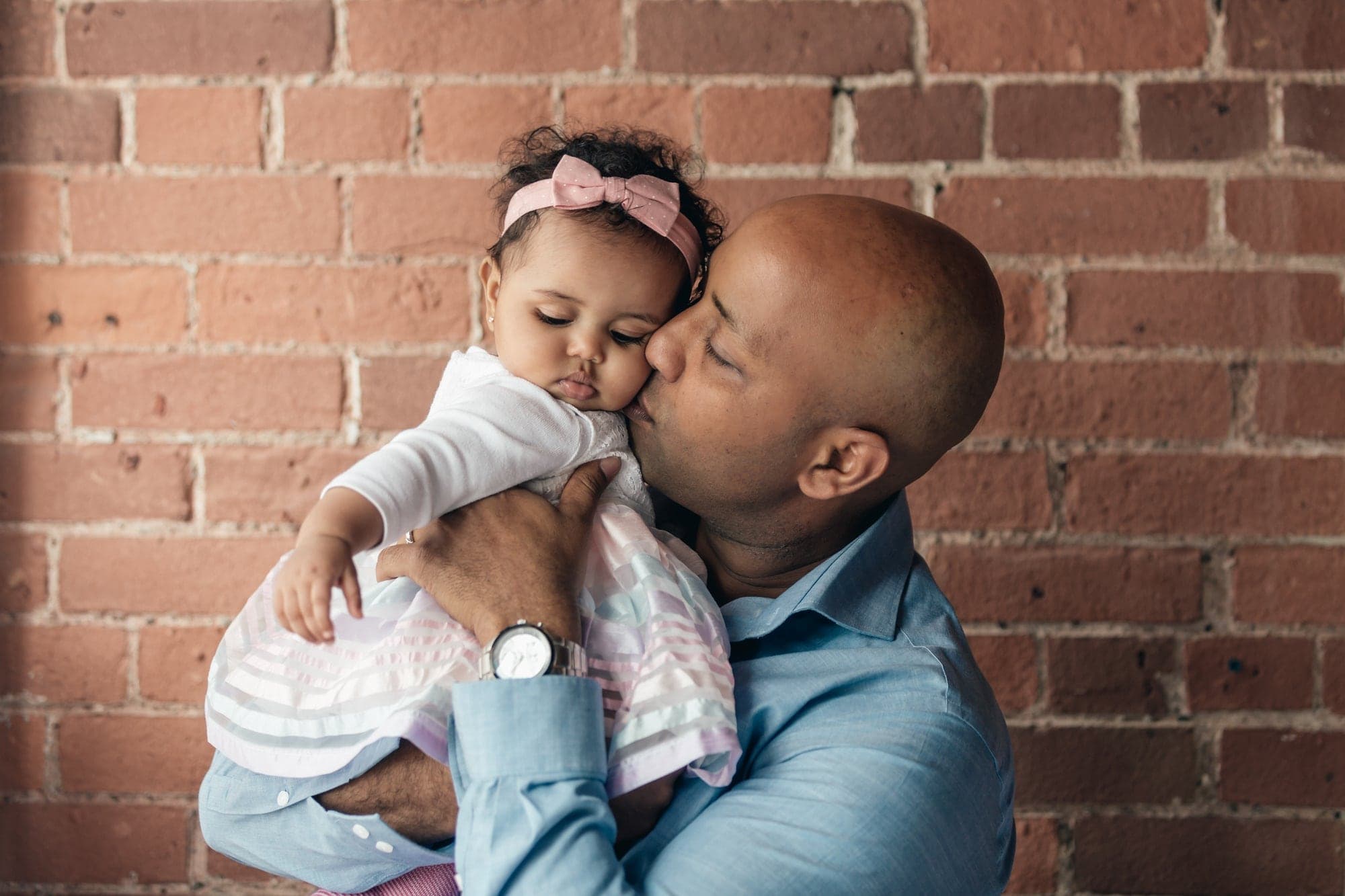 A father kissing his baby daughter's cheek against a brick wall